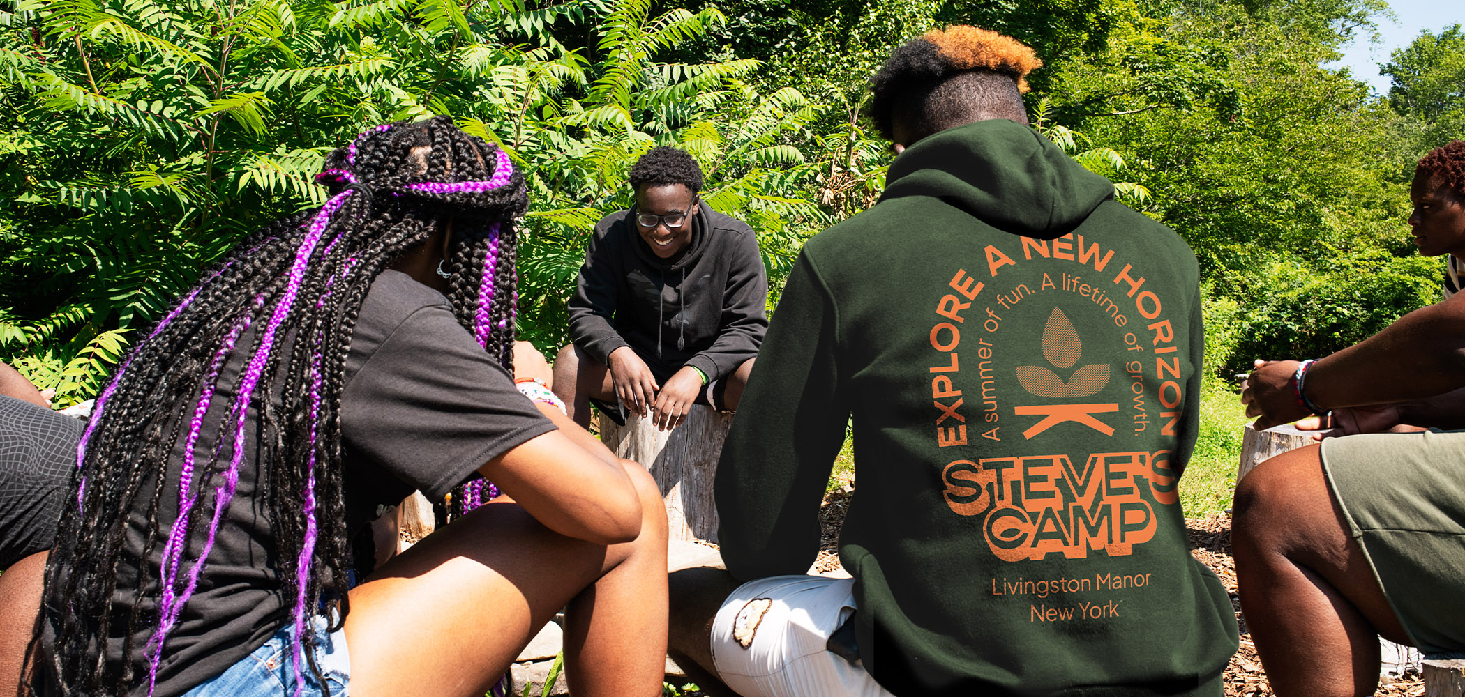 A group of smiling campers sit in a circle on logs.