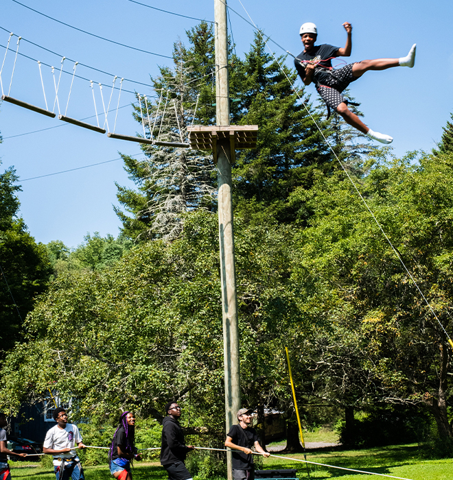 A boy swinging on a ropes course with other campers supporting him from below