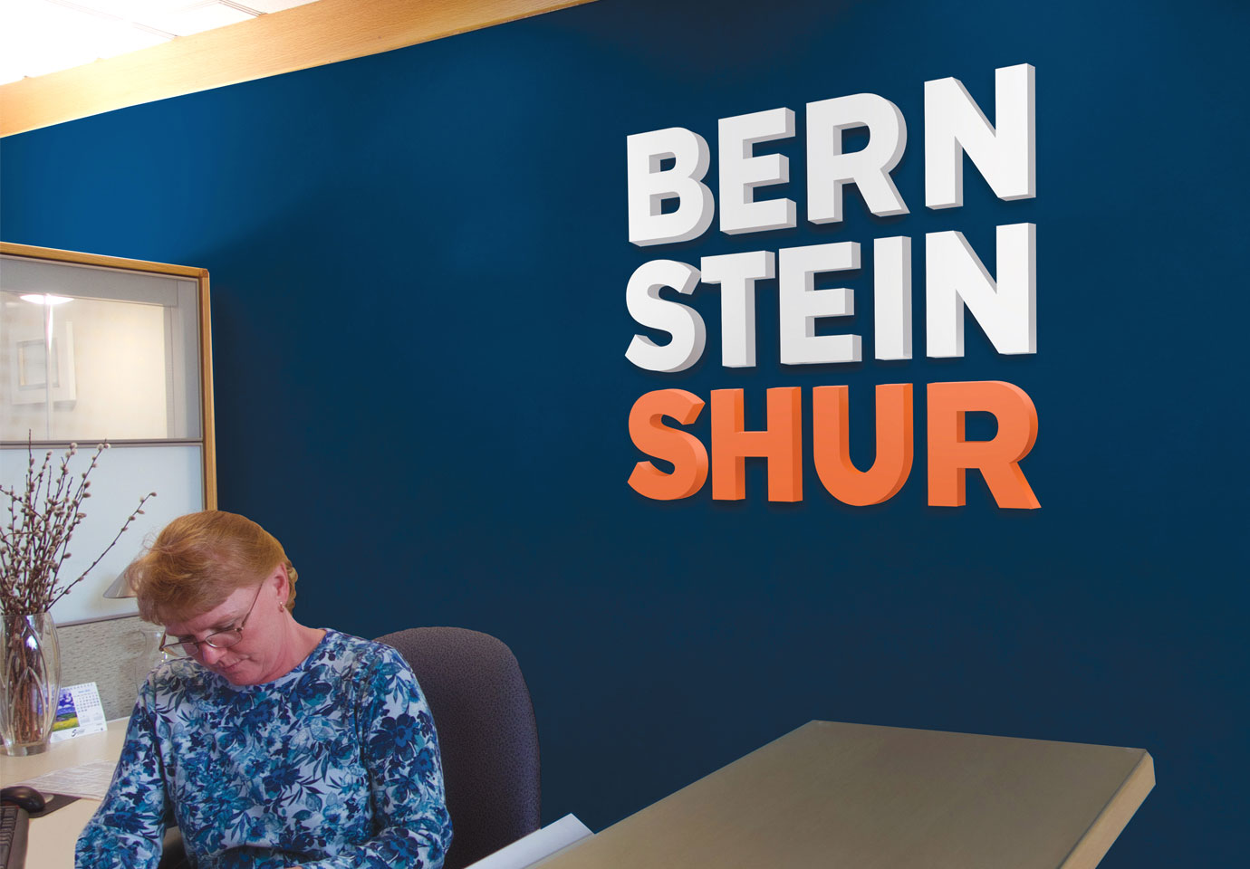 The Bernstein Shur logo on a dark blue wall in the company reception area. A person sits a the desk in the foreground.
