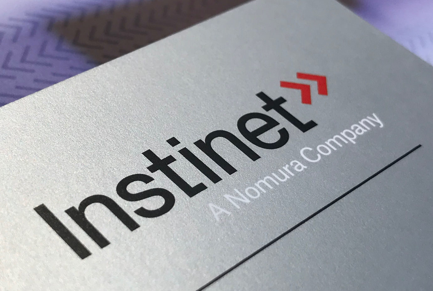 The Instinet logotype with black and white lettering on a silver surface and the red double chevron brand emblem.