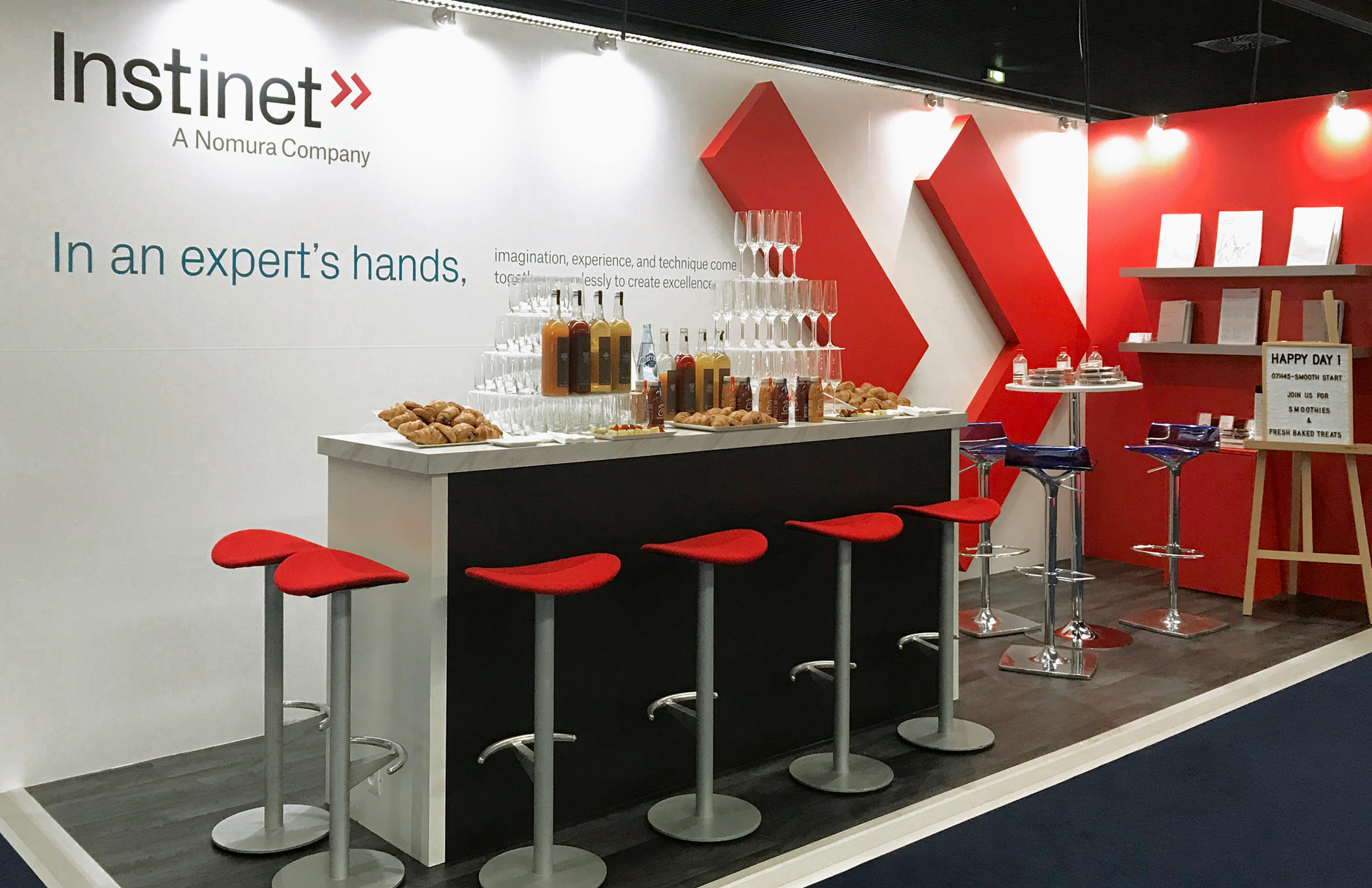 The Instinet reception booth at an industry conference with seating area in front of a giant, wall-mounted sculpture of the red, dual chevron brand emblem.