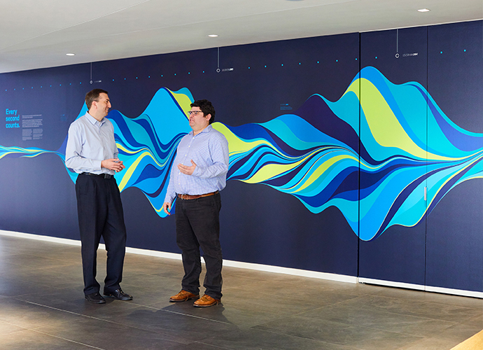Two KCG employees stand in front of a multi-colored wave pattern mural that depicts one second of market trading activity.