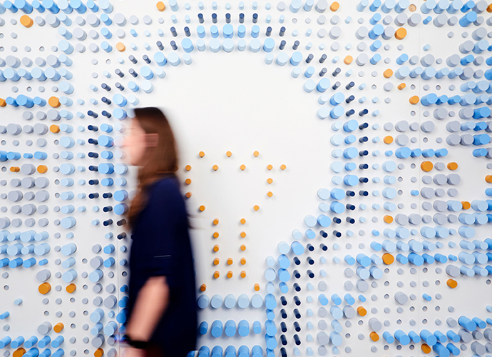 A figure walks in front of a  wall sculpture in the KCG office: the image of a light bulb formed from colored wooden dowels.