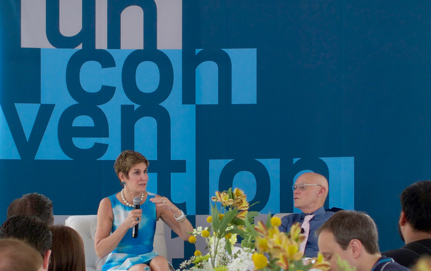 A seated presenter speaks to a group of attendees in front of a wall painted with a large KCG Un-convention event logo.