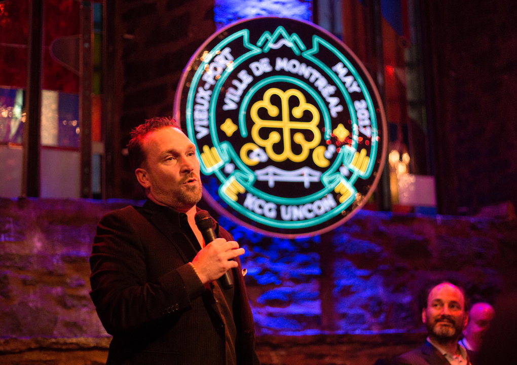 A speaker addresses attendees during an evening reception in front the neon sign logo for 2017 KCG Uncon client event.
