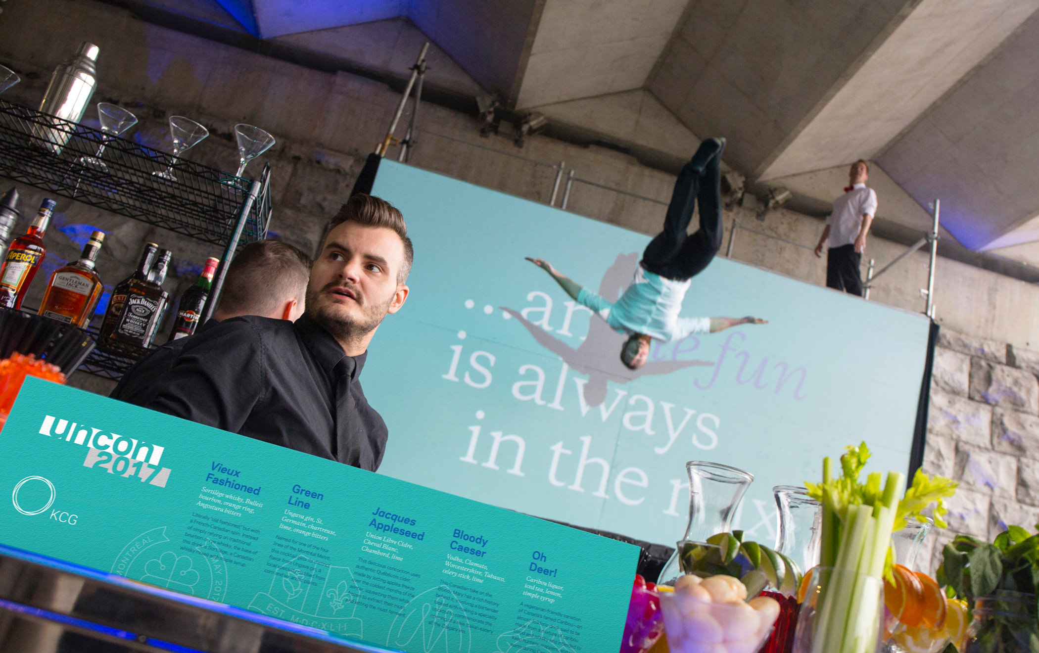 A bartender and cocktail menu sign at the 2017 KCG Uncon client event. A pair of acrobats perform in the background.