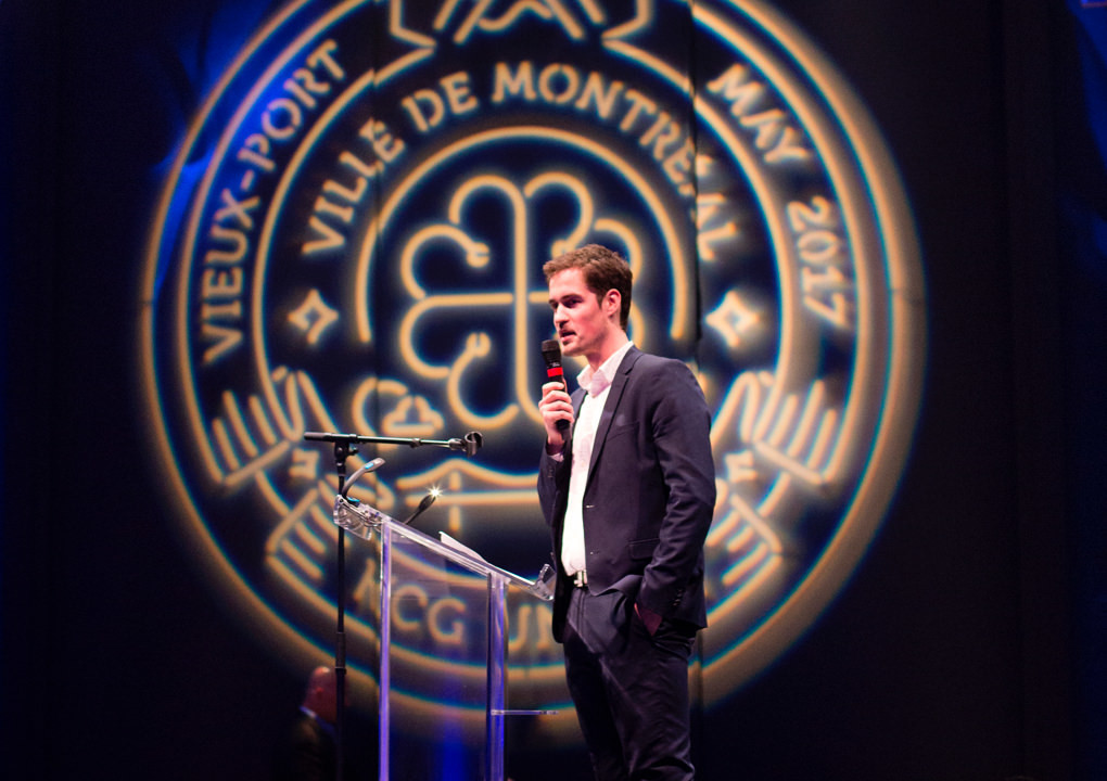 A single presenter at podium in front of a large neon sign logo for the 2017 KCG Uncon client event in Montreal.