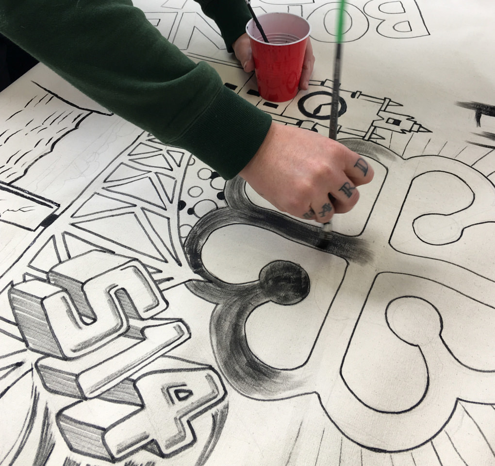 An artist painting and lettering a piece of white canvas to be cut and made into tote bags for the 2017 KCG Uncon event.
