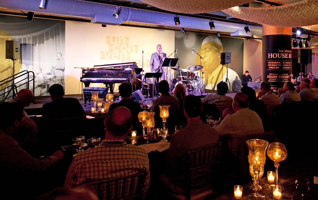 Branford Marsalis on the bandstand at a 2015 KCG Uncon client event reception with a large conference banner behind him.