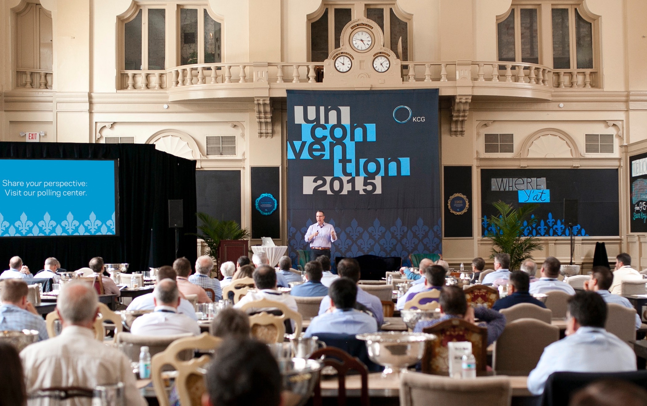 Presenter in a large hall in front of a giant conference banner speaking to a group of 2015 KCG Uncon attendees.
