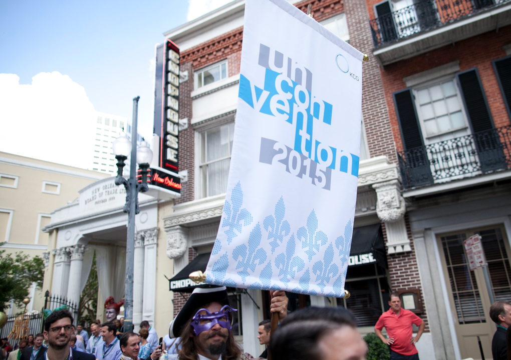 A 2015 KCG Uncon client event banner flies above attendees at a street parade in New Orleans.