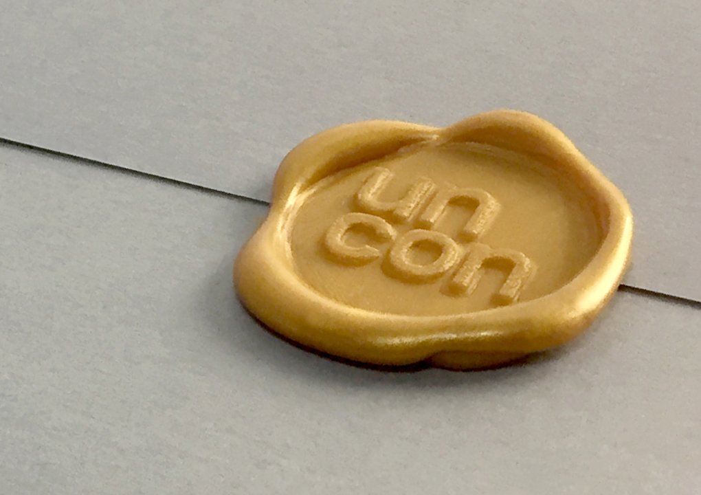 Close up a gold wax seal of the KCG Uncon client event logo on on a grey envelope.