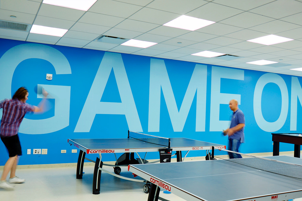 KCG employees play ping pong in front of a blue wall emblazoned with the phrase "Game on" in giant letters.