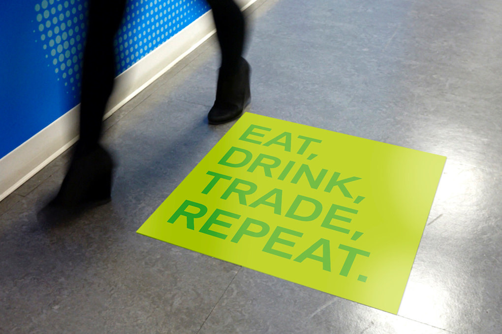 Blurred legs in motion walk over a floor sticker in the KCG cafeteria printed with the words "Eat, drink, trade, repeat."