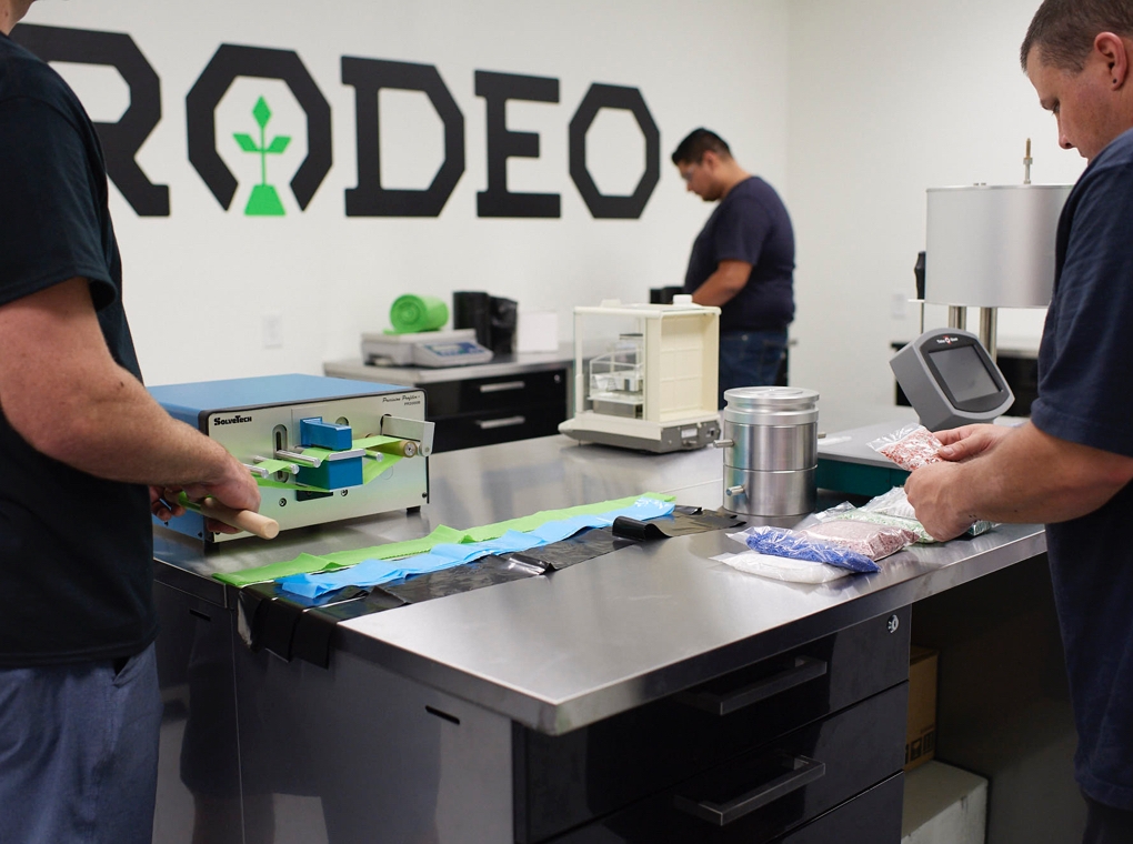 Three workers inspect products on tables in front of a wall painted with a large Rodeo Plastics logo.