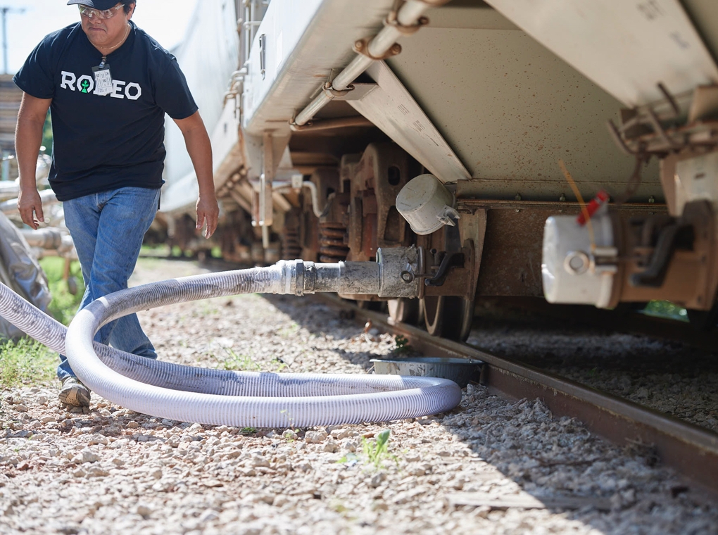 A worker in a Rodeo Plastics tee shirt attaching a hose to a railroad freight car tanker.
