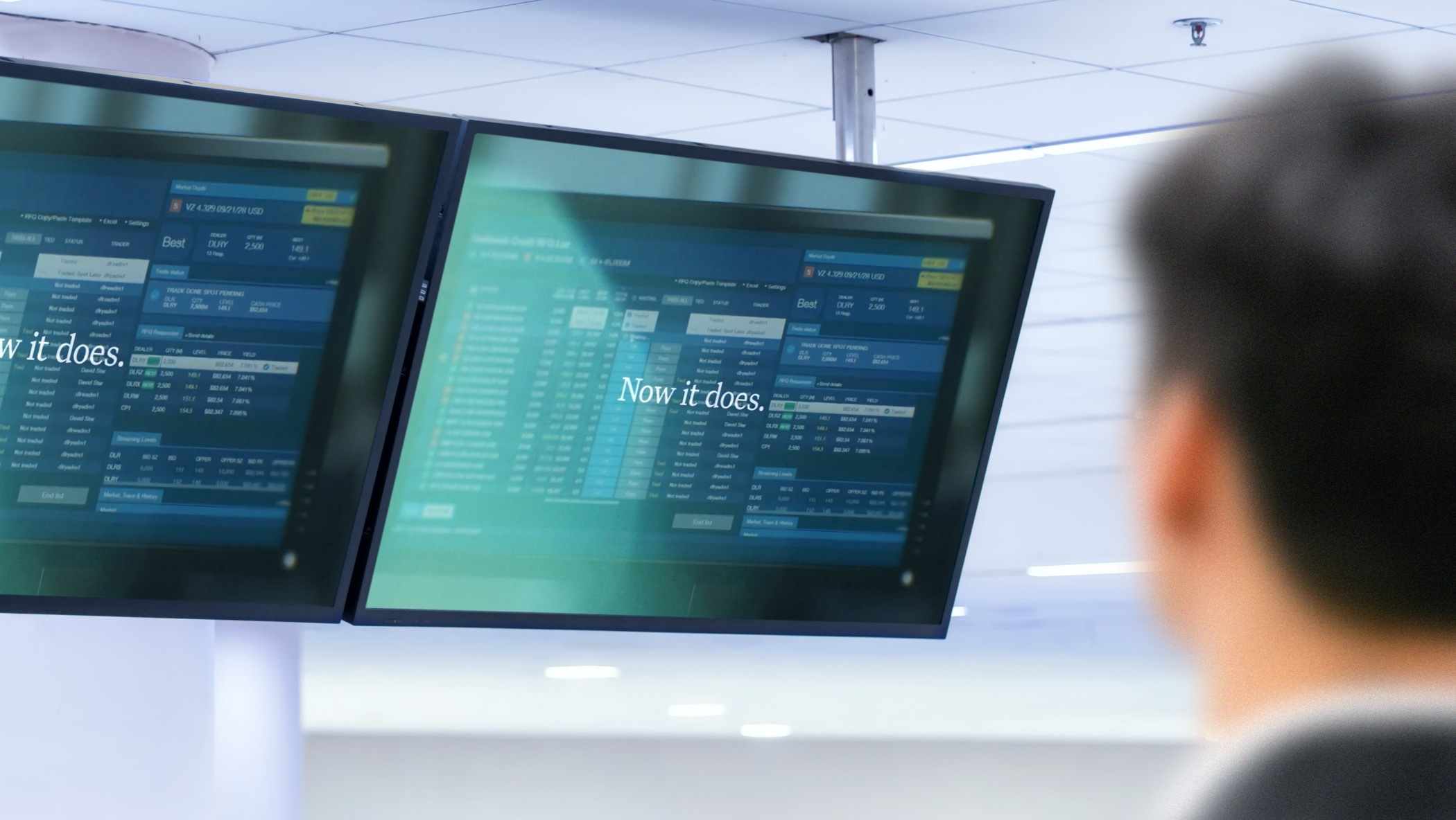 Overhead TV screens displaying a video still frame with the words “Now it does,” with a blurry figure in the foreground