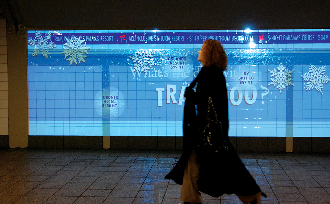 A figure walks in front of a large Travelzoo digital ad projected on a tile wall at Grand Central Station in New York City.