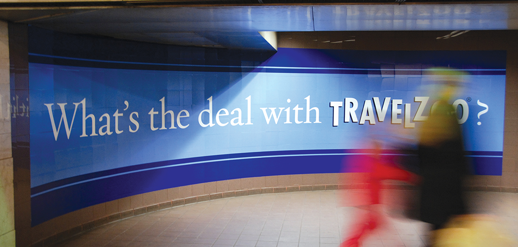 A mural-sized advertisement on a curved tile New York CIty subway wall that reads "What's the deal with Travelzoo?"