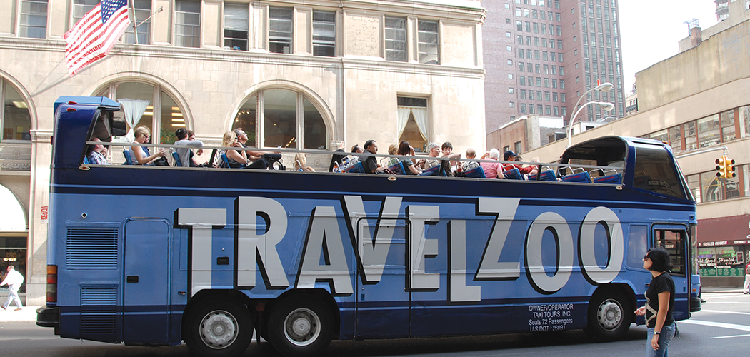 An open-top tourist bus on a city street with a giant Travelzoo logo painted on the side.