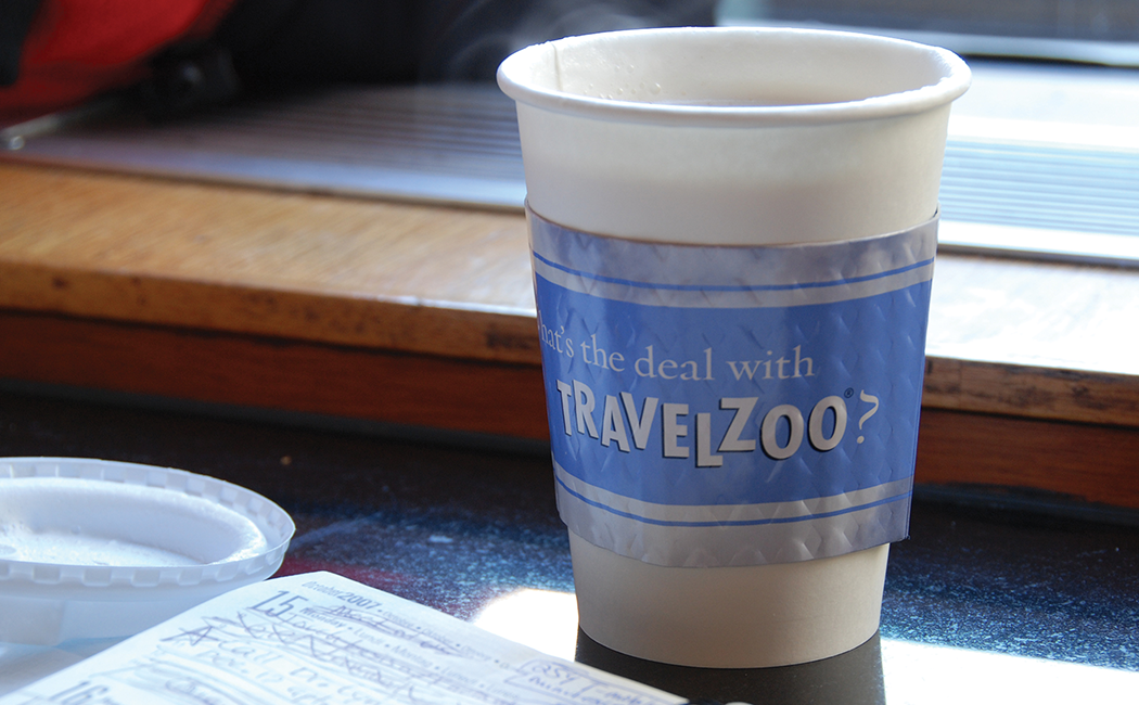 Close up of a to-go coffee cup with a cozy printed with the text "What's the deal with Travelzoo?"