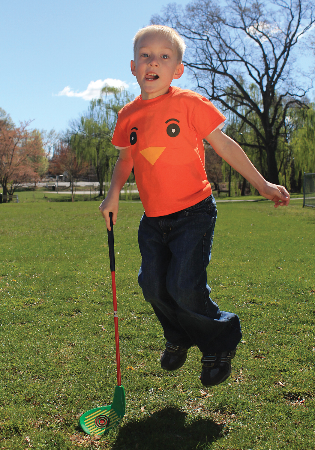 A young boy with a plastic golf club leaping up in his Urban Golf Academy "birdie" mascot T shirt.