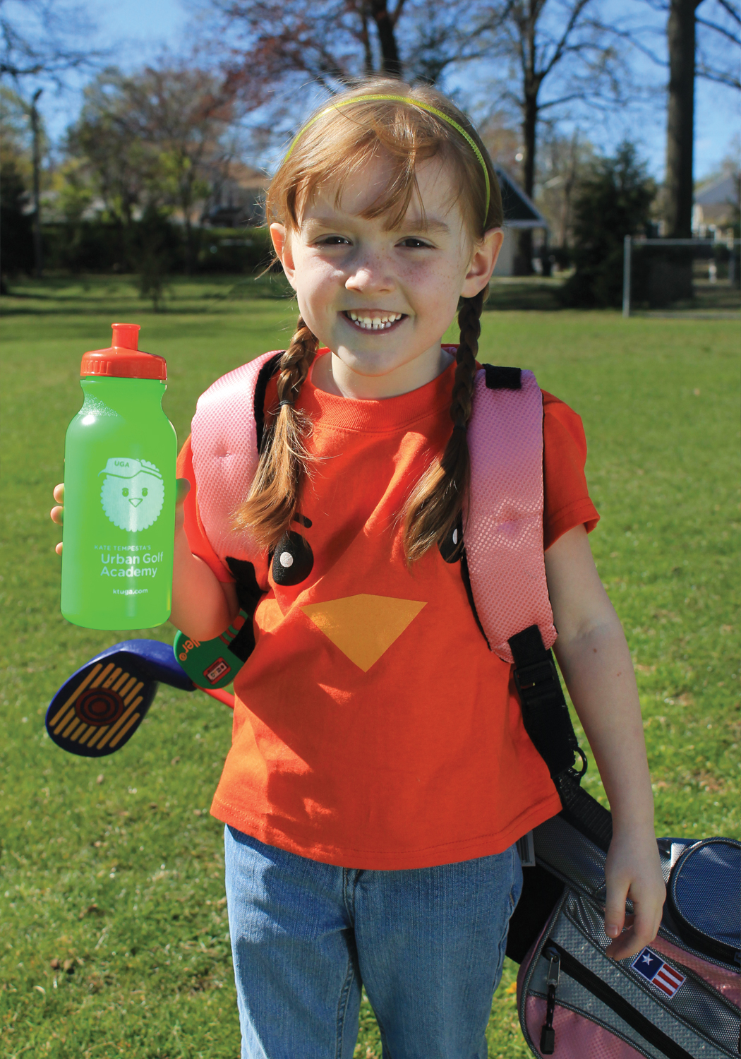 A young girl carrying a golf bag with an Urban Golf Academy water bottle and "birdie" mascot T shirt.