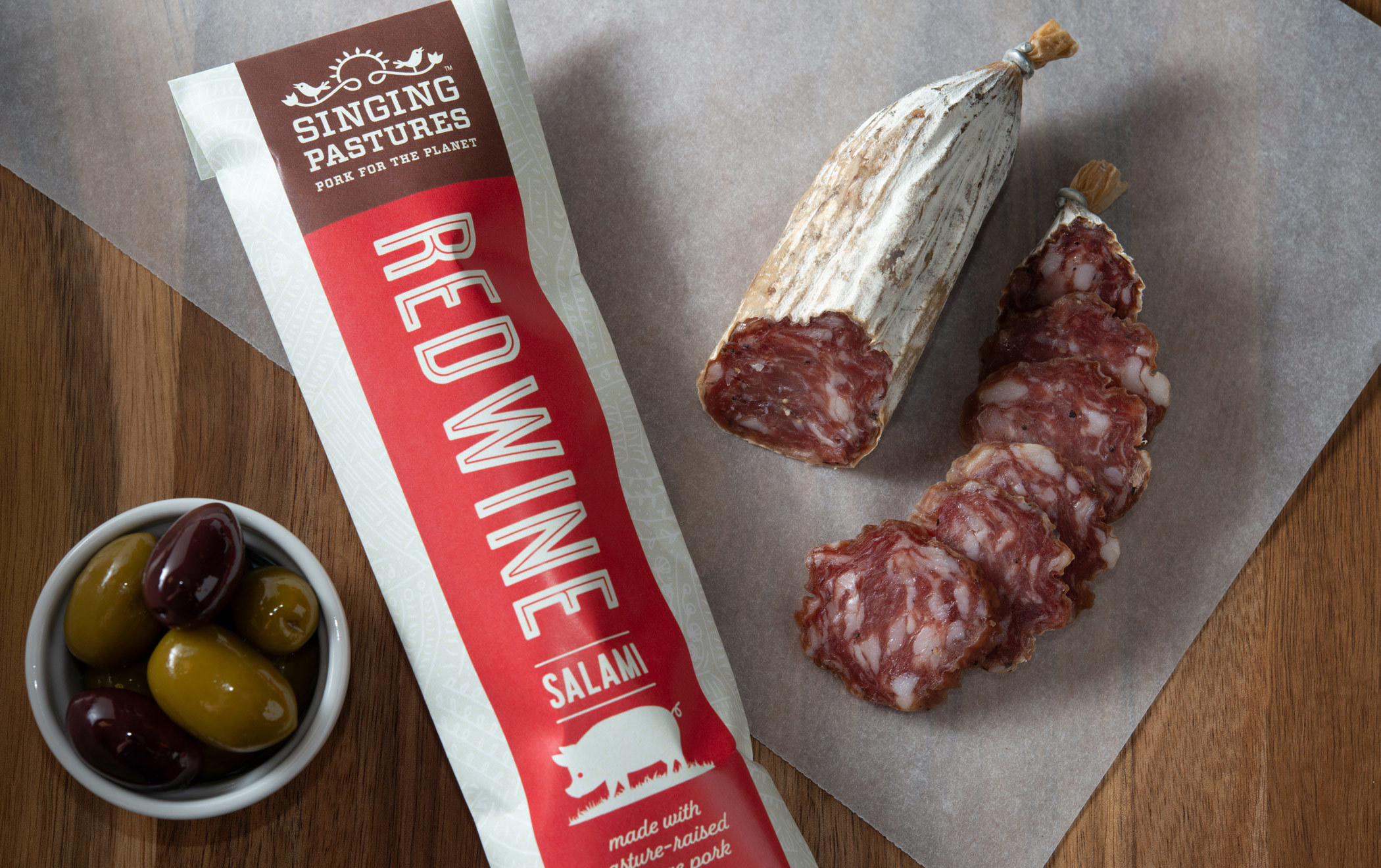 A red wine flavored Singing Pastures Roam Stick package next to a sliced sausage and small dish of olives.