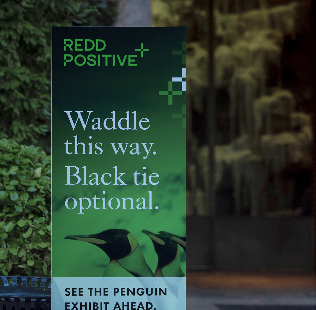 A large vertical outdoor sign directs guests at the REDD Positive Cocktails and Conservation launch event to the penguin exhibit with the headline, Waddle this way. Black tie optional.